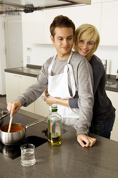 Junger Mann beim Kochen und eine junge Frau  die ihn von hinten in der Küche umarmt.