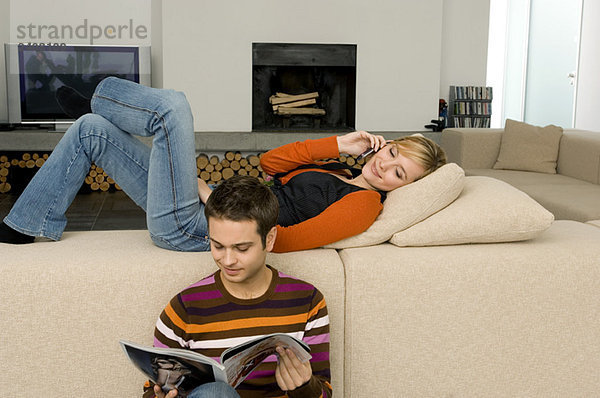 Junger Mann liest eine Zeitschrift mit einer jungen Frau  die ein Handy hinter sich hat.