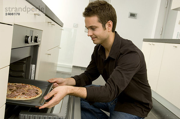 Junger Mann stellt ein Tablett mit Pizza in den Ofen und lächelt.