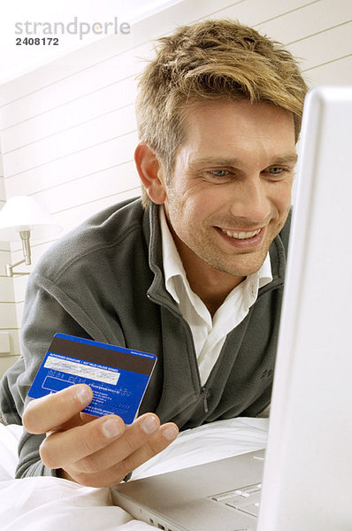 Mittlerer erwachsener Mann  der an einem Laptop arbeitet und eine Kreditkarte hält.