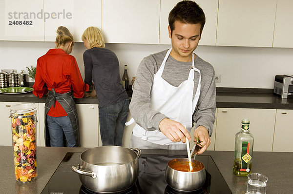 Junger Mann beim Kochen mit zwei jungen Frauen  die hinter ihm stehen.