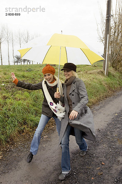 Zwei junge Frauen  die sich unter einem Regenschirm verstecken und auf einem Feldweg spazieren gehen.