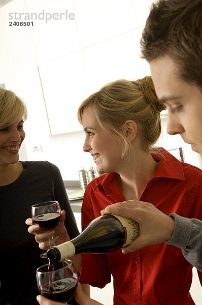 Junger Mann gießt Wein in ein Weinglas mit zwei jungen Frauen  die neben ihm lächeln.
