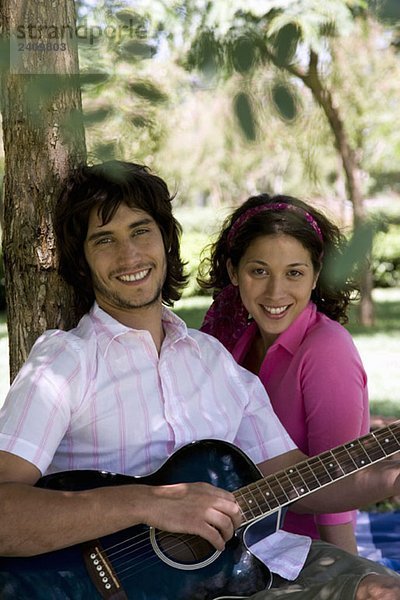 Ein junges Paar sitzt unter einem Baum und spielt Gitarre.