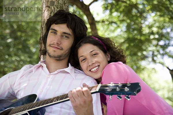 Ein junges Paar sitzt unter einem Baum und spielt Gitarre.