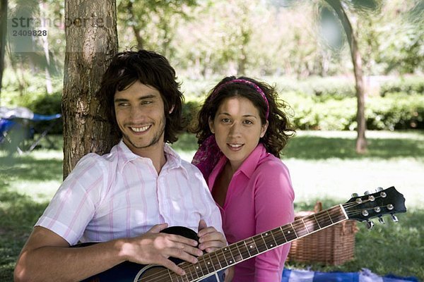 Ein junger Mann und eine junge Frau sitzen unter einem Baum mit einer Gitarre.