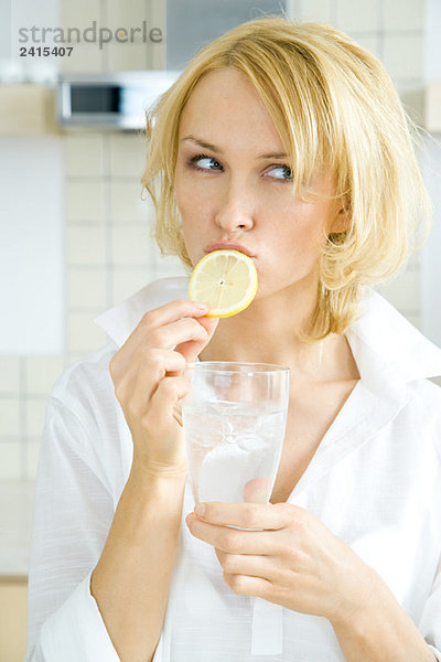Junge Frau beißt in eine Zitronenscheibe  schaut weg  hält ein Glas Wasser.