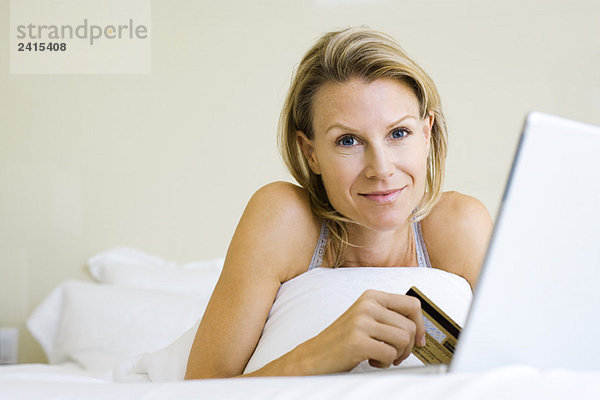 Frau auf dem Bett liegend  mit Kreditkarte und Laptop  lächelnd vor der Kamera