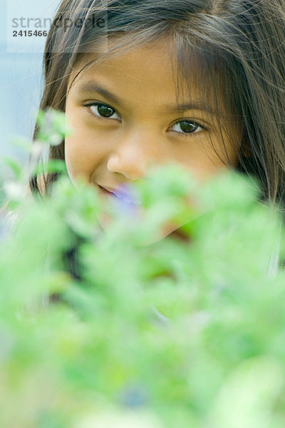 Mädchen lächelt in die Kamera  schaut über die Pflanze im verschwommenen Vordergrund