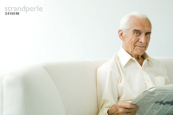 Älterer Mann auf dem Sofa sitzend  Zeitung haltend  lächelnd vor der Kamera
