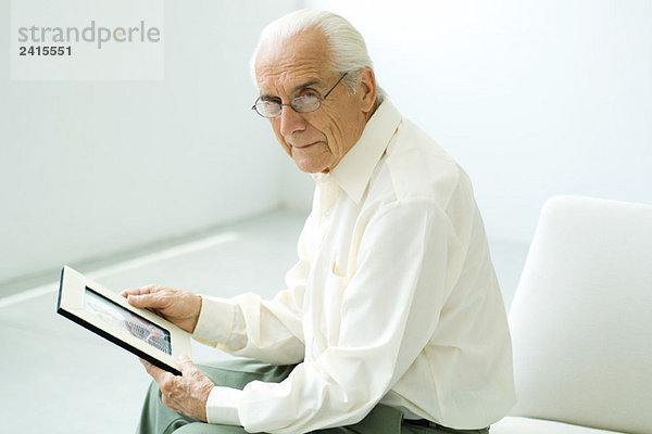 Älterer Mann sitzt  hält ein Foto  schaut in die Kamera.