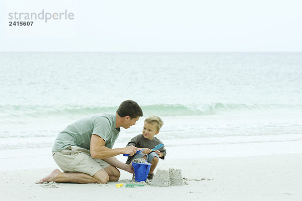 Vater und Sohn bauen Sandburg am Strand  lächeln sich an.