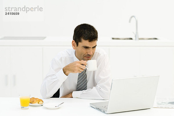 Geschäftsmann am Küchentisch sitzend  mit Blick auf den Laptop  Kaffee trinken