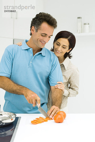 Paar steht zusammen in der Küche  Mann schneidet Tomate  beide lächelnd