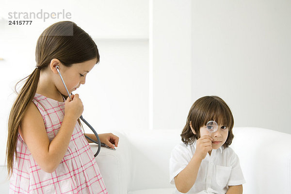 Kleiner Junge schaut durch Lupe auf die Kamera  Mädchen hört Stethoskop