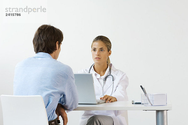 Ärztin sitzt dem männlichen Patienten am Schreibtisch gegenüber  mit Laptop-Computer
