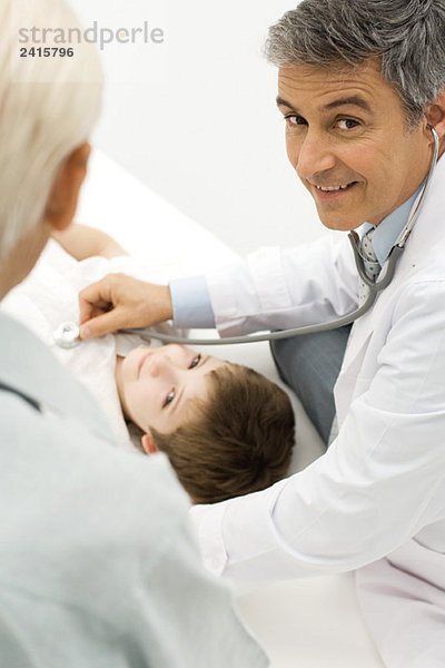 Arzt hört auf das Herz des jungen Patienten  lächelt über die Schulter vor der Kamera