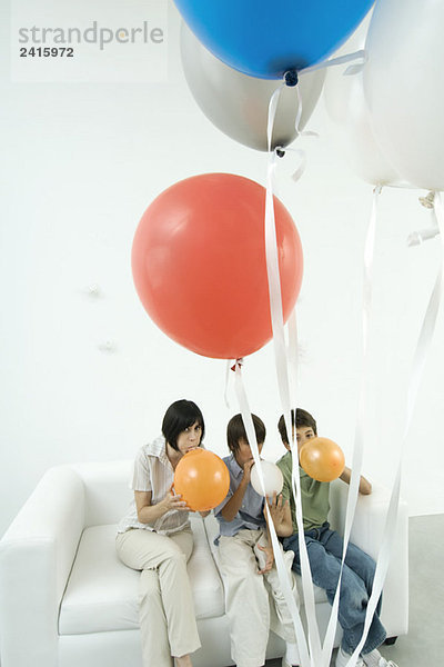 Mutter und zwei Söhne auf dem Sofa sitzend  Luftballons aufblasend