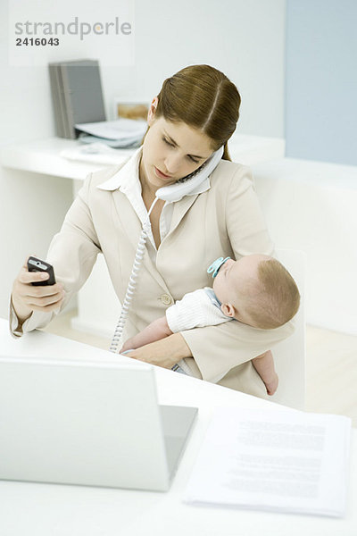 Junge berufstätige Mutter hält Baby  spricht am Festnetztelefon und schaut aufs Handy