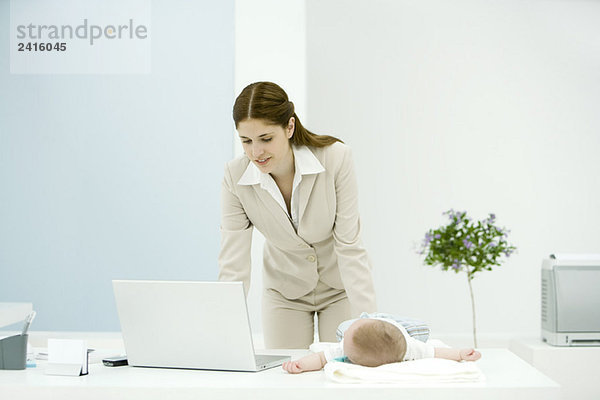 Junge berufstätige Mutter mit Laptop  Baby auf dem Schreibtisch liegend