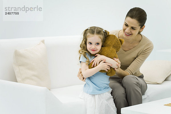 Kleines Mädchen mit Plüschbär  Frau auf der Couch sitzend  kleines Mädchen sanft umarmend
