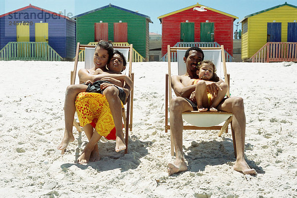 Familie am Strand  Eltern sitzen in Liegestühlen und halten Kinder auf dem Schoß.