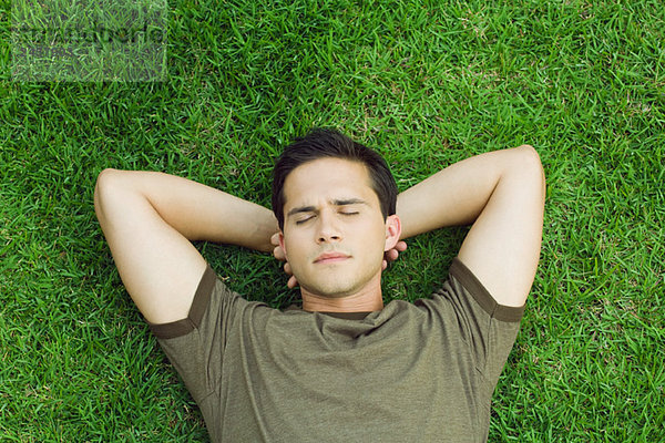 Junger Mann auf Gras liegend  Hände hinter dem Kopf  Augen geschlossen  Hochwinkelansicht