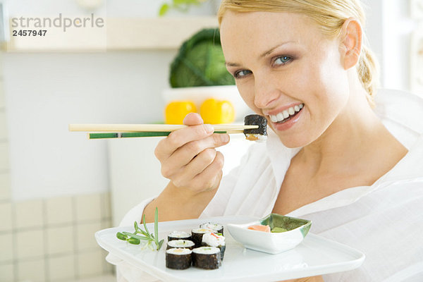 Frau isst Maki-Sushi mit Stäbchen und lächelt in die Kamera.