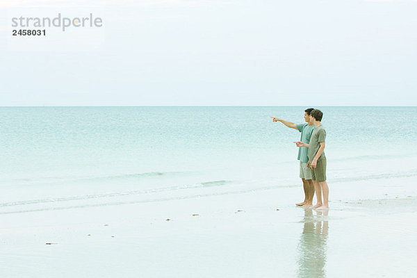 Mann und Teenager am Strand stehend  mit Blick auf die Aussicht  Mann zeigt