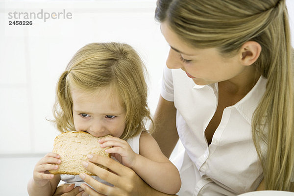 Kleines Mädchen sitzt auf dem Schoß ihrer Mutter und isst eine Scheibe Brot.