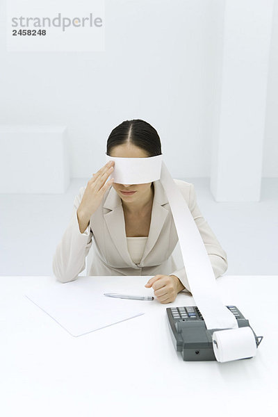 Frau am Schreibtisch sitzend  Klebeband von einer Addiermaschine um die Augen gewickelt  Gesicht berührend