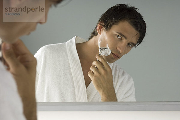 Der Mann schaut sich im Spiegel an  rasiert sich  trägt einen Bademantel.