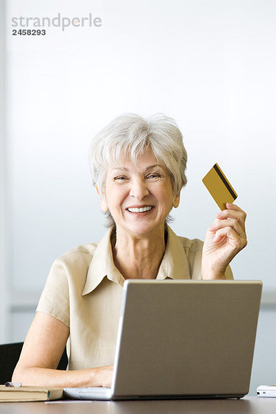 Seniorin am Schreibtisch sitzend  mit Kreditkarte  mit Laptop  lächelnd vor der Kamera
