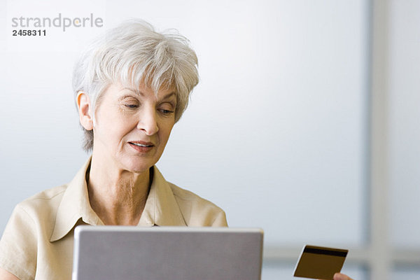 Senior Frau schaut auf Kreditkarte  sitzt hinter dem Laptop