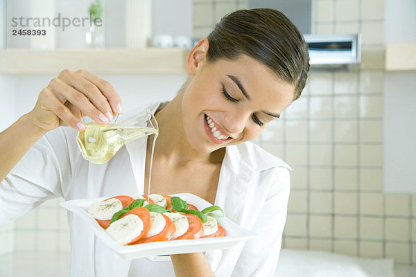 Junge Frau gießt Olivenöl über Tomaten-Mozzarella-Salat  lächelnd