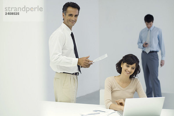 Berufstätige im Büro  Mann steht und hält Dokument  Frau sitzt vor dem Laptop