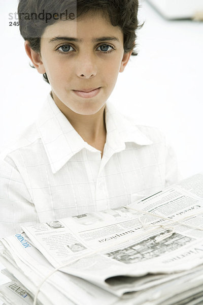 Junge mit einem Stapel Zeitungen  lächelnd vor der Kamera.