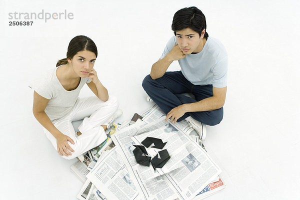 Junges Paar sitzt neben einem Stapel Zeitungen mit Recyclingsymbol und blickt in die Kamera.