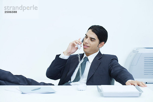 Junger Geschäftsmann sitzt mit erhobenen Füßen am Schreibtisch und telefoniert auf dem Festnetz.
