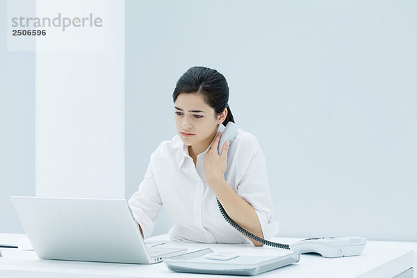 Junge berufstätige Frau am Schreibtisch sitzend  mit Laptop  Festnetztelefon an der Brust haltend
