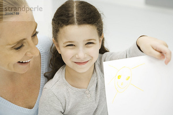 Mädchen hält sich hoch und zeigt die Zeichnung der Sonne  Mutter lächelt hinter ihrer Tochter.