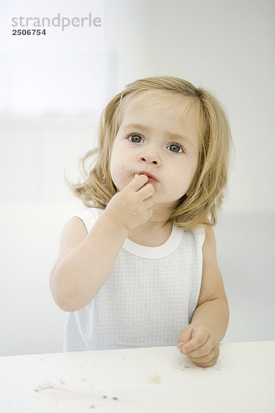 Kleines Mädchen sitzt am unordentlichen Tisch und wischt sich eine Krume von der Lippe.