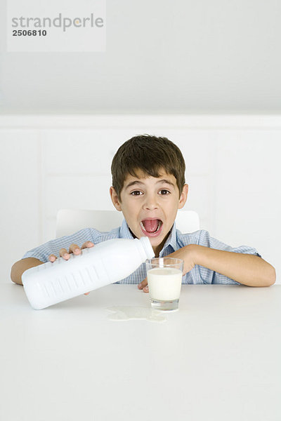Junge gießt Milch ins Glas  Mund offen  verschüttete Milch auf dem Tisch
