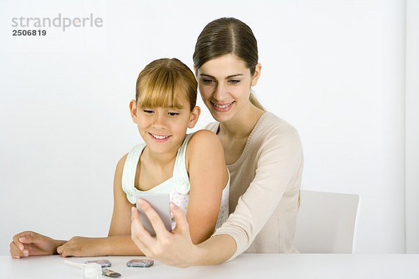 Mädchen sitzt auf dem Schoß einer jungen Frau und schaut zusammen in den Handspiegel.
