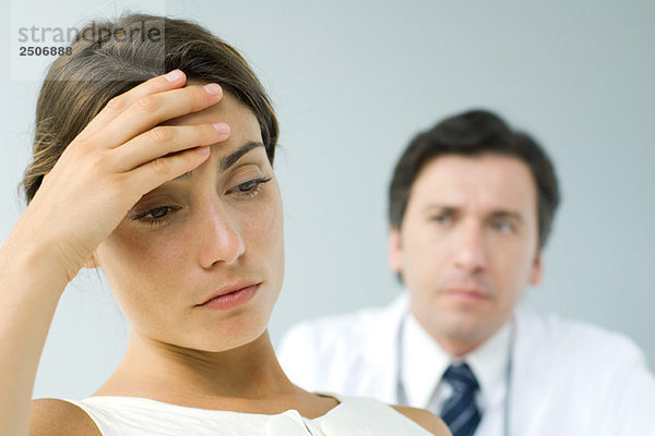 Frau hält Kopf  schaut nach unten  Arzt im Hintergrund