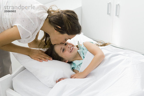 Mädchen im Bett liegend  Mutter bückt sich  um ihre Stirn zu küssen.