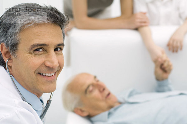 Doktor lächelt in die Kamera  älterer Mann liegt auf der Couch im Hintergrund