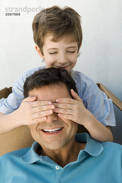 Junge mit den Händen über den Augen seines Vaters  beide lächelnd  die Augen des Jungen geschlossen.