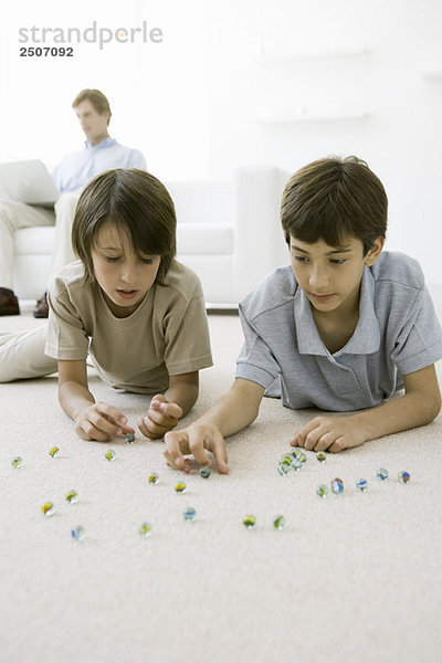 Jungen liegen auf dem Boden und spielen mit Murmeln  Vater sitzt auf dem Sofa mit Laptop im Hintergrund.