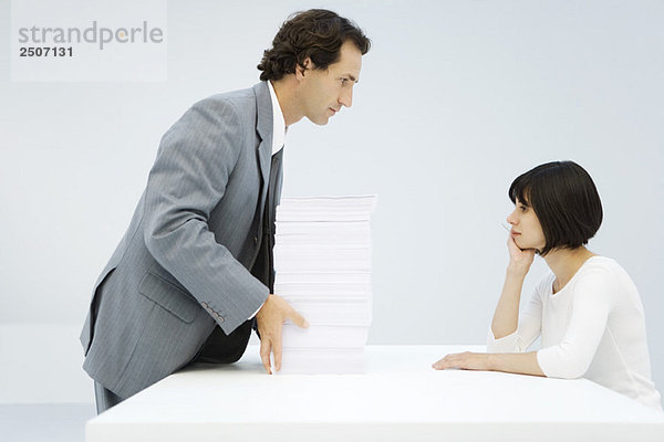 Geschäftsmann schiebt einen hohen Stapel Papier über einen Tisch zu einer Frau  die sich auf ihren Ellbogen stützt.